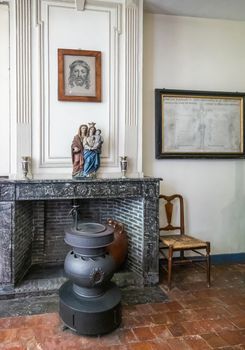 Bruges, Flanders, Belgium -  June 17, 2019: Living room of house in Beguinage comes with Catholic religious art, a sober black stove, a simple brown chair and a red tiled uneven floor.