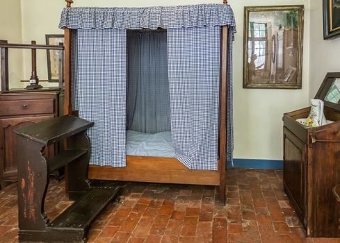 Bruges, Flanders, Belgium -  June 17, 2019: Bed room of house in Beguinage comes with a bed with curtains, a sober brown praying stool, simple furniture, washing set, and a red tiled uneven floor.