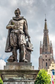 Bruges, Flanders, Belgium -  June 17, 2019: Simon Stevin statue with spire of OLV Cathedral in back against rainy cloudscape. Some green foliage. Pigeon on his head.