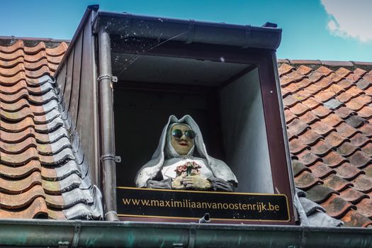 Bruges, Flanders, Belgium -  June 17, 2019: Closeup of nun with sun glasses sculpture set in roof window of restaurant Maximiliaan van Oostenrijk. Red roof, blue sky.