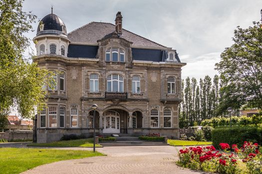 Knokke-Heist, Flanders, Belgium -  June 18, 2019: Kursaalstraat. Historic brown-gray building of Provincial Fishermen School, now defunct, in its park under cloudscape. Green foliage and red flowers.