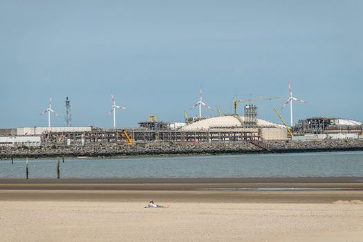 Zeebrugge, Flanders, Belgium -  June 18, 2019: Closeup of LNG terminal in port of Zeebrugge under blue sky as seen from beach in Knokke-Heist. Lone sun bather on sand.