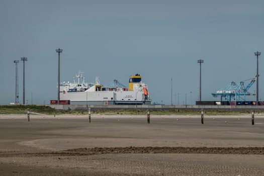 Zeebrugge, Flanders, Belgium -  June 18, 2019: Ferry and container ship with Container cranes in port of Zeebrugge, as seen from beach in Knokke-Heist. Gray-blue sky, wet sand.