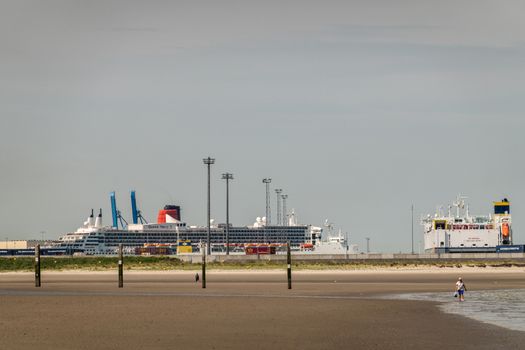 Zeebrugge, Flanders, Belgium -  June 18, 2019: Queen Mary 2 Cruise Ship in port of Zeebrugg as seen from beach in Knokke-Heist under gray-blue sky. Wet sand in front. Walkers.