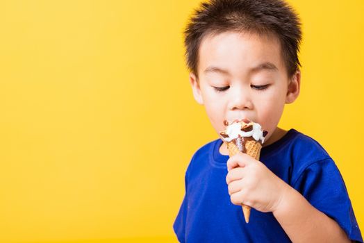 Happy portrait Asian child or kid cute little boy attractive laugh smile playing holds and eating sweet chocolate ice cream waffle cone, studio shot isolated on yellow background, summer concept