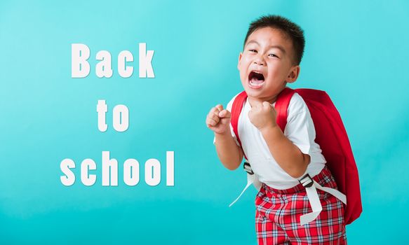 Back to school. Portrait happy Asian cute little child boy in uniform smile raise hands up glad when go back to school, isolated blue background. Kid from preschool kindergarten with school backpack