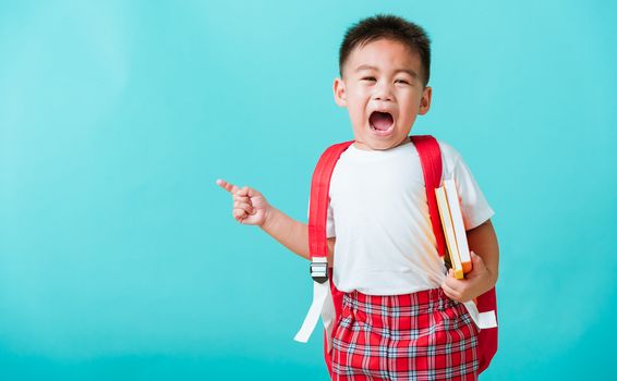 Back to school concept. Portrait Asian happy funny cute little child boy smile hug books and point finger to side away space, isolated blue background. Kid from preschool kindergarten with school bag