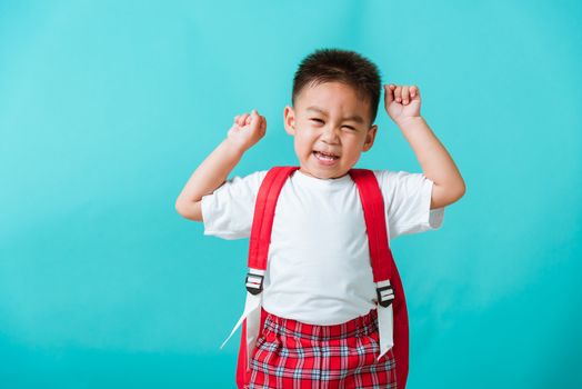 Back to school. Portrait happy Asian cute little child boy in uniform smile raise hands up glad when go back to school, isolated blue background. Kid from preschool kindergarten with school backpack