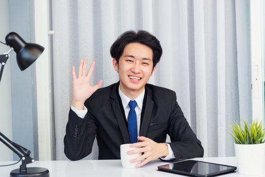 Work from home, Asian young businessman video conference call or facetime he smiling looking to camera sitting on desk using smart digital tablet computer raise hand to say hello at home office