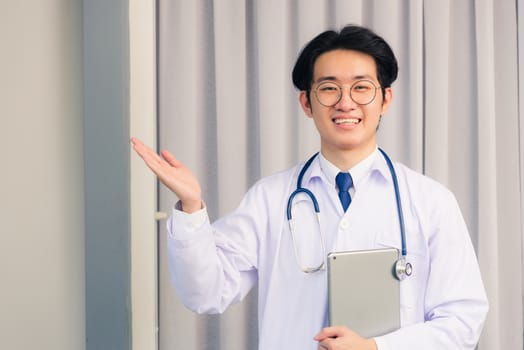 Portrait closeup of Happy Asian young doctor handsome man smiling in uniform and stethoscope neck strap holding smart digital tablet on hand and showing hand to side away, healthcare medicine concept