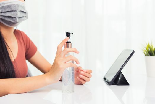 Asian Business young woman working from home office he quarantines disease coronavirus or COVID-19 wearing a protective mask and cleaning hands with sanitizer gel on front tablet computer