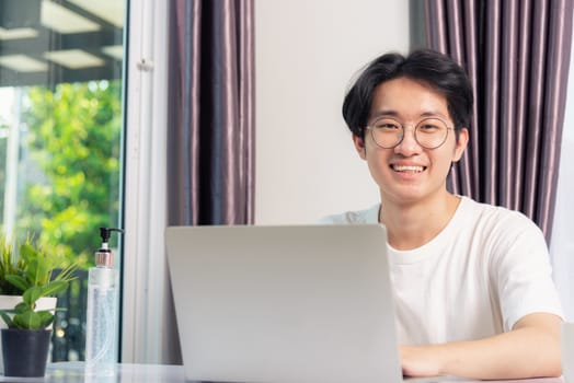 Happy Asian young business handsome man smile work from home office wear glasses and t-shirt comfortable working with laptop computer on desk, Thai person freelance man worker at home