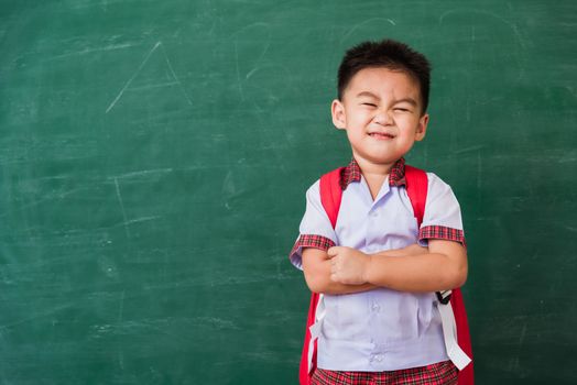 Back to School. Happy Asian funny cute little child boy from kindergarten in student uniform with school bag stand smile crossed arm on green school blackboard, First time to school education concept