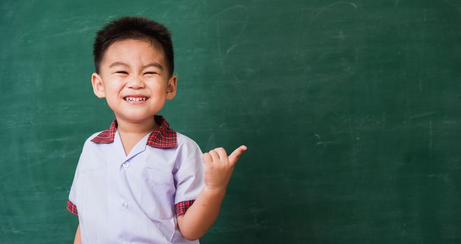 Back to School. Happy Asian funny cute little child boy from kindergarten in student uniform smiling point finger to side away space on green school blackboard, First time to school education concept