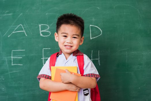 Back to School. Happy Asian funny cute little child boy kindergarten in student uniform with school bag and books smile show finger thumb up on green school blackboard, First time to school education