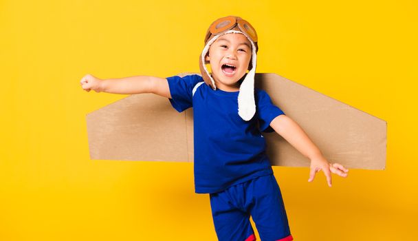 Happy Asian handsome funny child or kid little boy smile wear pilot hat playing and goggles with toy cardboard airplane wings flying, studio shot isolated yellow background, Startup freedom concept