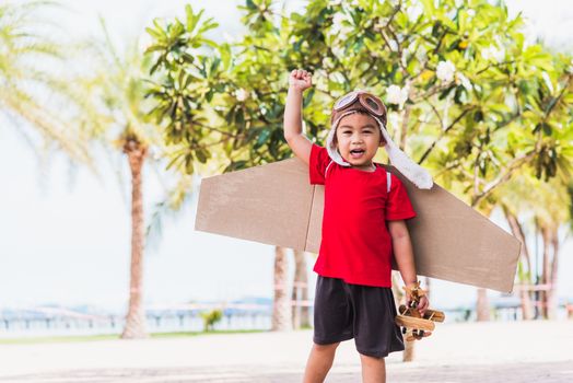 Happy Asian funny child or kid little boy smile wear pilot hat and goggles play toy cardboard airplane wing flying against summer sky cloud on trees garden background, Startup freedom concept