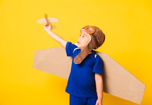 Happy Asian handsome funny child or kid little boy smile wear pilot hat play and goggles with toy cardboard airplane wings fly hold plane toy, studio shot isolated yellow background, Startup freedom