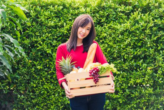 Portrait of Asian beautiful young woman farmer standing she smile and holding full fresh food raw vegetables fruit in a wood box in her hands on green leaves background