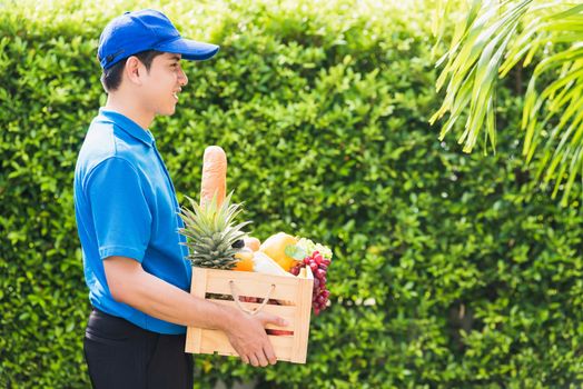 Asian man farmer wears delivery uniform he holding full fresh vegetables and fruits in crate wood box in hands ready give to customer harvest organic food on the garden place green leaves background