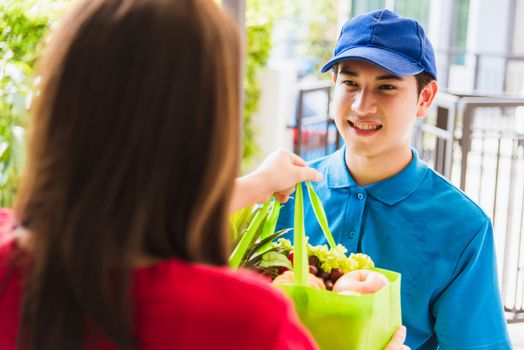 Asian young delivery man in uniform making grocery service giving fresh vegetables and fruits and food in green cloth bag to woman customer at door house after pandemic coronavirus, Back to new normal