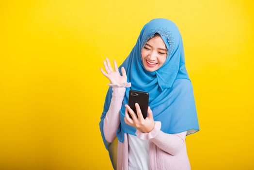 Asian Muslim Arab, Portrait of happy beautiful young woman Islam religious wear veil hijab funny smile she selfie or video call mobile smart phone raise hand say hello isolated on yellow background