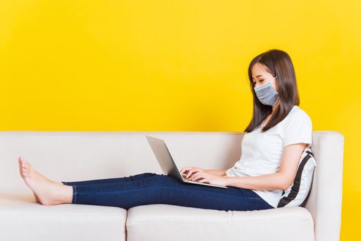 Portrait Asian of beautiful young woman sitting on sofa wearing medical face mask protective she work from home with laptop computer during Coronavirus studio shot isolated on yellow background