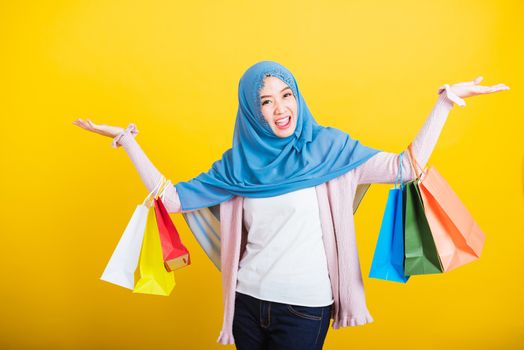 Asian Muslim Arab, Portrait of happy beautiful young woman Islam religious wear veil hijab funny smile she holding colorful shopping bags so glad shopping bags hand raise in studio isolated on yellow