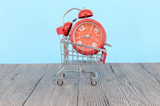 Shopping cart and classic alarm clock on wooden surface. Sale time buy mall market shop consumer concept. Selective focus.