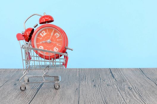 Shopping cart and classic alarm clock on wooden surface. Sale time buy mall market shop consumer concept. Selective focus.