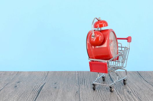 Shopping cart and classic alarm clock on wooden surface. Sale time buy mall market shop consumer concept. Selective focus.