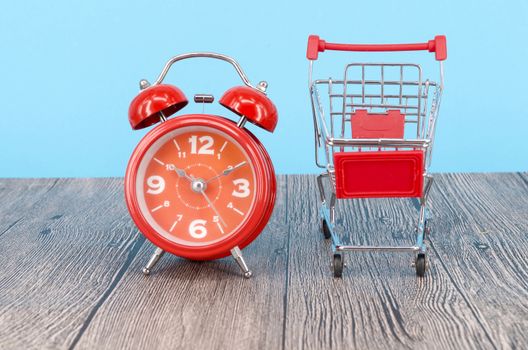 Shopping cart and classic alarm clock on wooden surface. Sale time buy mall market shop consumer concept. Selective focus.