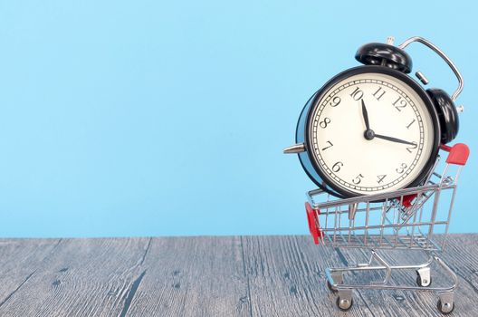 Shopping cart and classic alarm clock on wooden surface. Sale time buy mall market shop consumer concept. Selective focus.