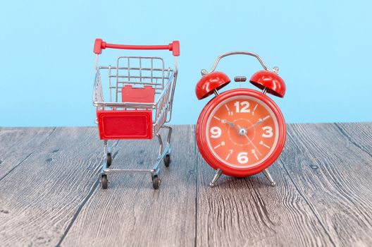 Shopping cart and classic alarm clock on wooden surface. Sale time buy mall market shop consumer concept. Selective focus.