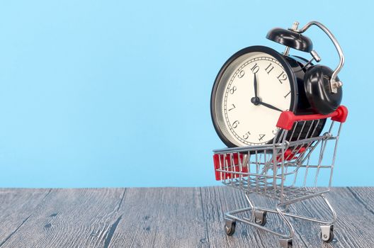 Shopping cart and classic alarm clock on wooden surface. Sale time buy mall market shop consumer concept. Selective focus.