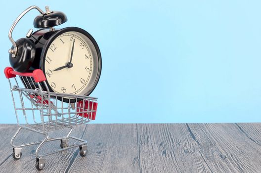 Shopping cart and classic alarm clock on wooden surface. Sale time buy mall market shop consumer concept. Selective focus.
