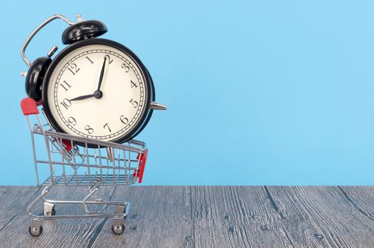 Shopping cart and classic alarm clock on wooden surface. Sale time buy mall market shop consumer concept. Selective focus.