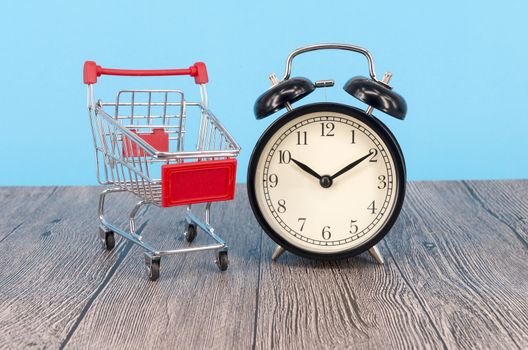 Shopping cart and classic alarm clock on wooden surface. Sale time buy mall market shop consumer concept. Selective focus.