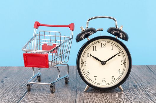 Shopping cart and classic alarm clock on wooden surface. Sale time buy mall market shop consumer concept. Selective focus.