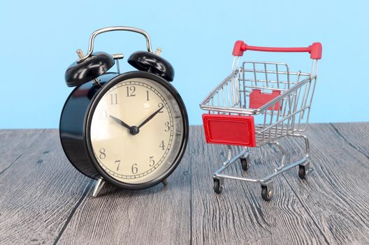 Shopping cart and classic alarm clock on wooden surface. Sale time buy mall market shop consumer concept. Selective focus.