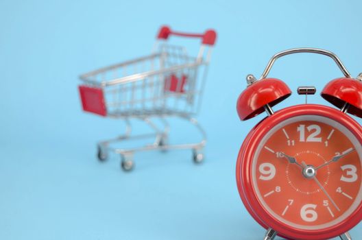 Shopping cart and classic alarm clock on blue background. Sale time market shop consumer concept. Selective focus.