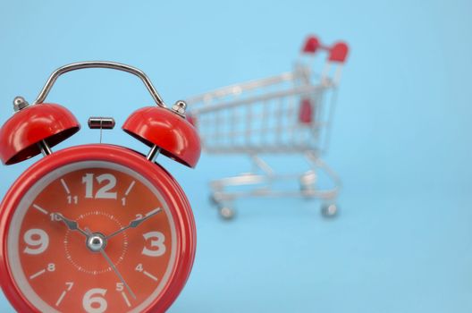 Shopping cart and classic alarm clock on blue background. Sale time market shop consumer concept. Selective focus.