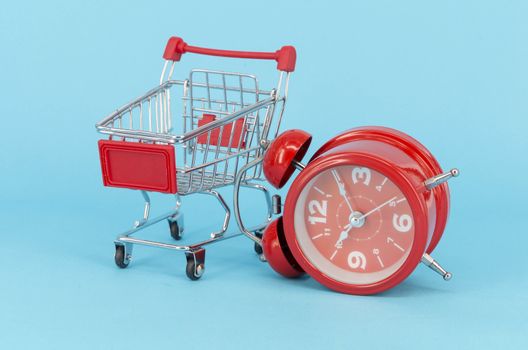 Shopping cart and classic alarm clock on blue background. Sale time market shop consumer concept. Selective focus.