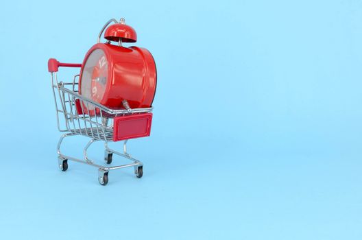 Shopping cart and classic alarm clock on blue background. Sale time market shop consumer concept. Selective focus.