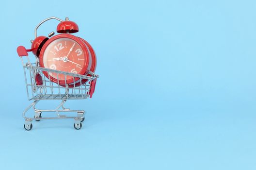 Shopping cart and classic alarm clock on blue background. Sale time market shop consumer concept. Selective focus.