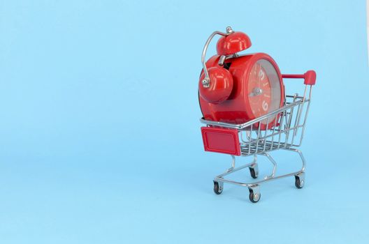 Shopping cart and classic alarm clock on blue background. Sale time market shop consumer concept. Selective focus.