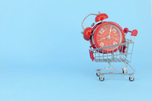 Shopping cart and classic alarm clock on blue background. Sale time market shop consumer concept. Selective focus.