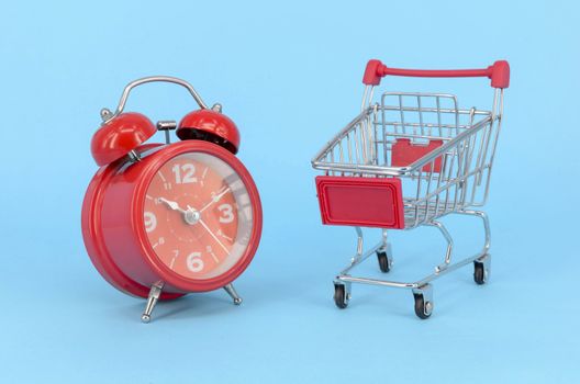 Shopping cart and classic alarm clock on blue background. Sale time market shop consumer concept. Selective focus.