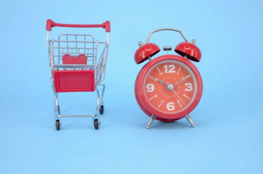 Shopping cart and classic alarm clock on blue background. Sale time market shop consumer concept. Selective focus.