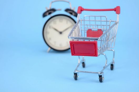 Shopping cart and classic alarm clock on blue background. Sale time market shop consumer concept. Selective focus.